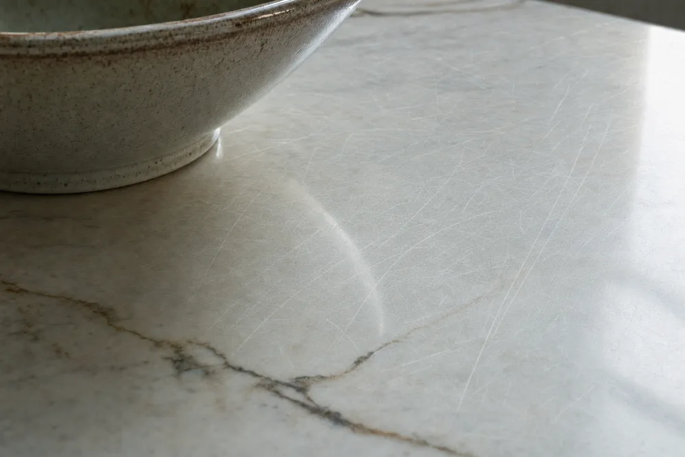 Fine scratches catching the light on pale polished marble beside a ceramic bowl