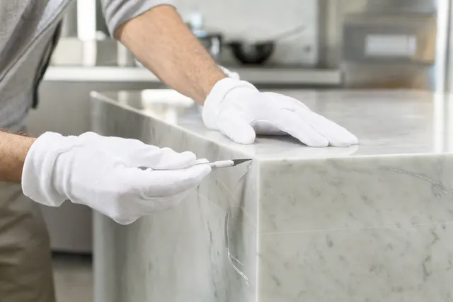 White-gloved hands trimming clear protection film flush along the edge of a marble island benchtop