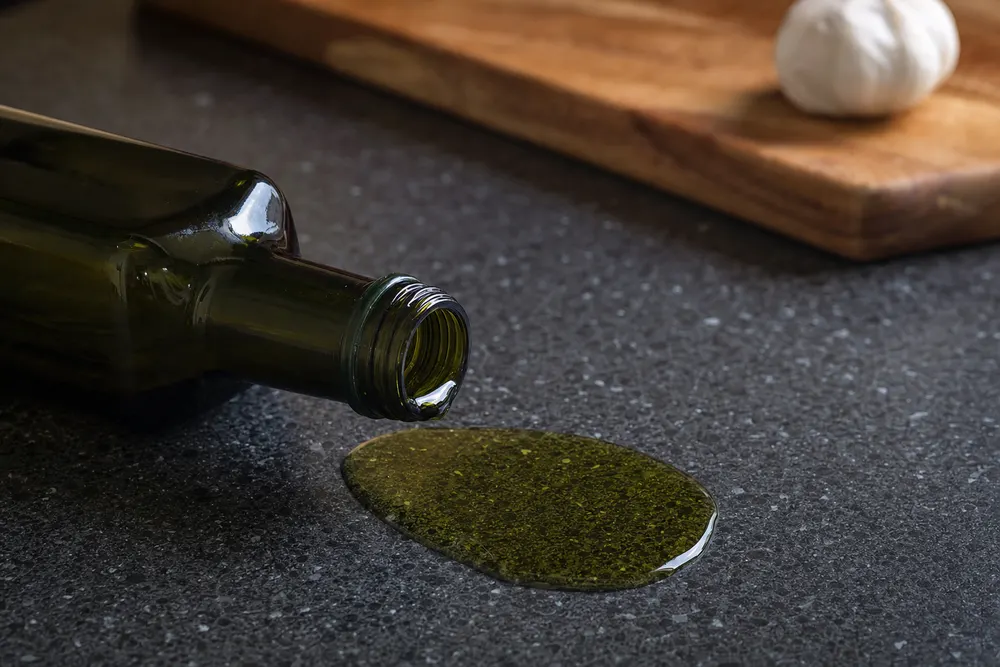 A tipped olive oil bottle with oil pooling on a speckled charcoal granite benchtop