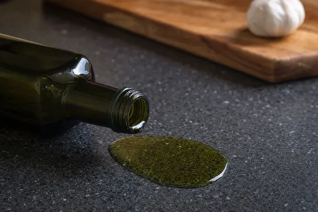 A tipped olive oil bottle with oil pooling on a speckled charcoal granite benchtop